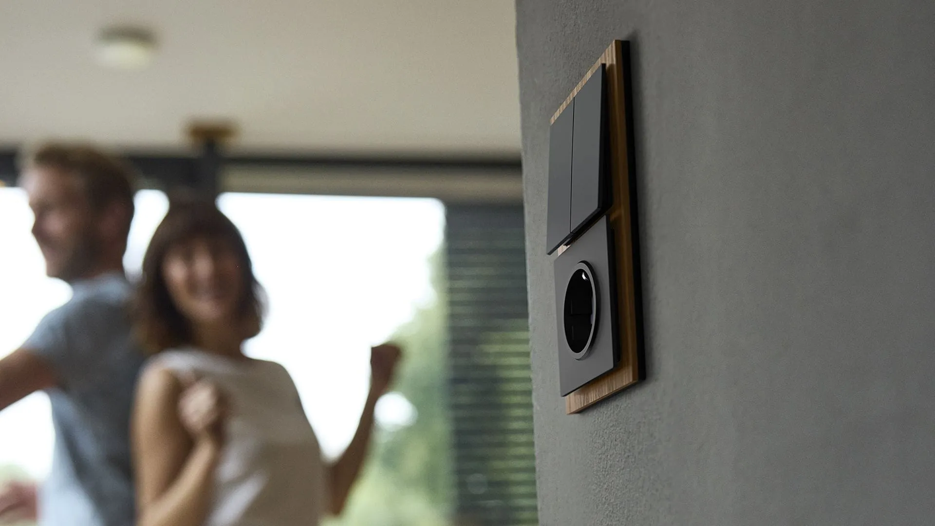 Busch-art linear Busch-Jaeger switch and socket mounted on a wall, with a couple in the blurred background