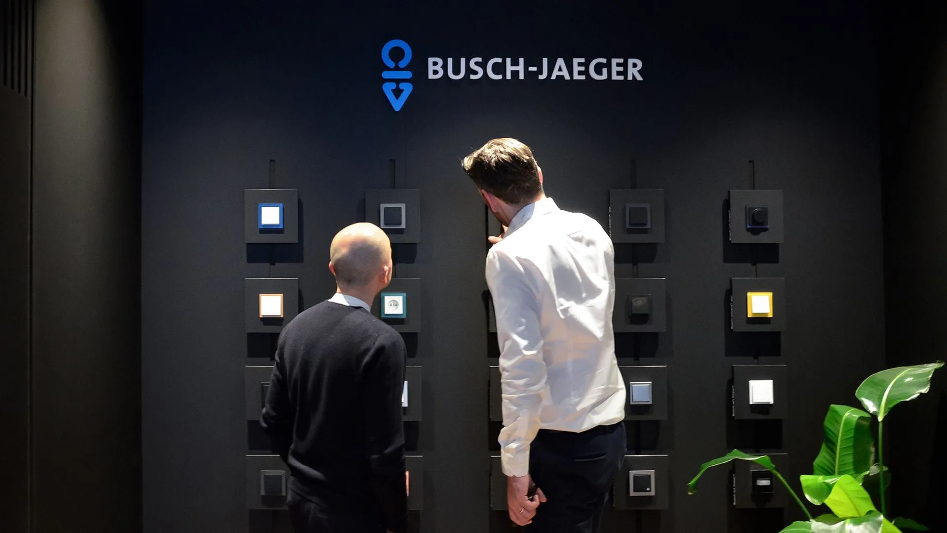 Two visitors exploring the Busch-Jaeger showroom wall displaying Busch-art linear switches at a trade fair