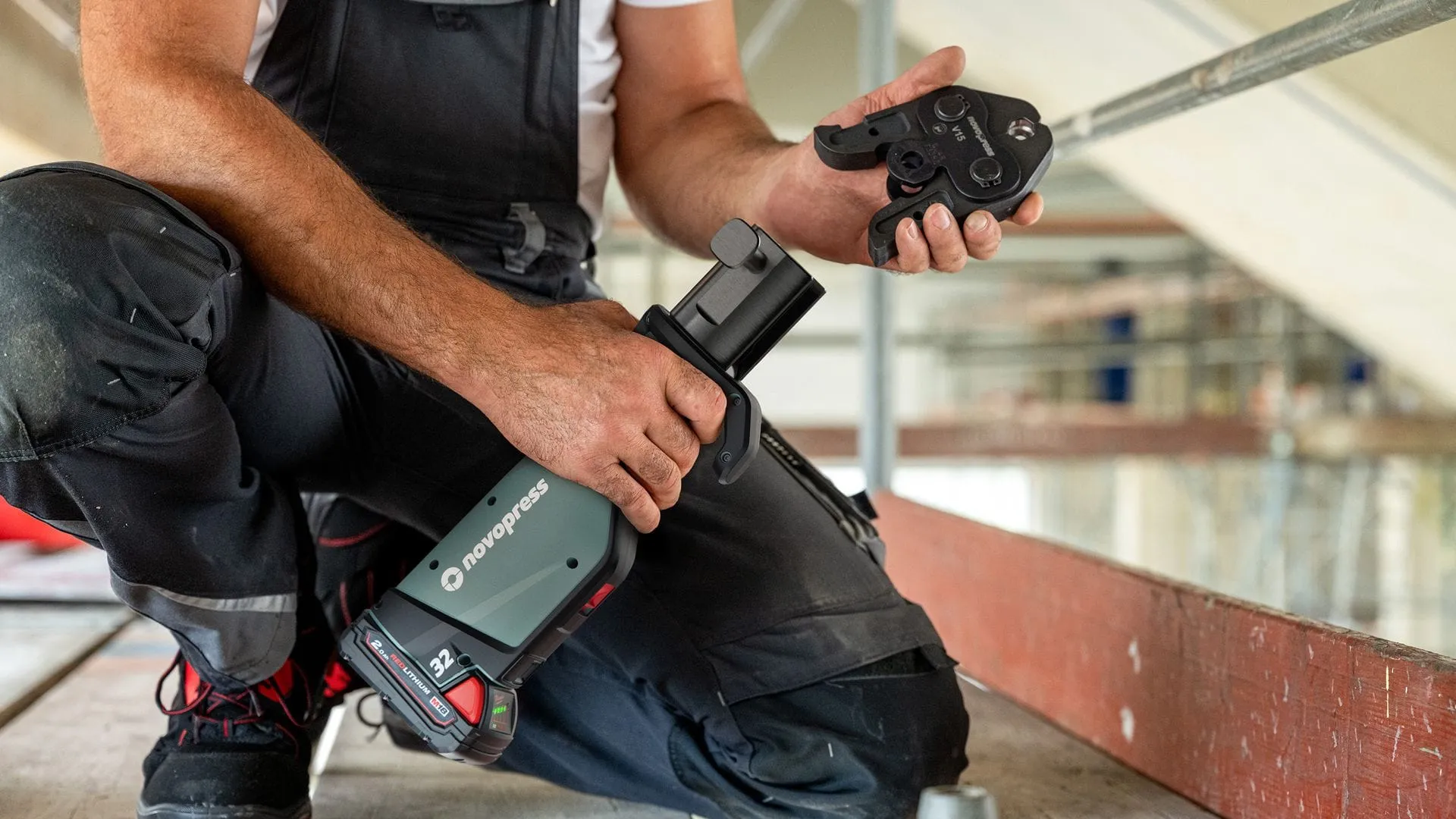 Installateur kauert auf einer Baustelle und hält die NOVOPRESS 32 Pressmaschine und einen abmontierten Pressbacken